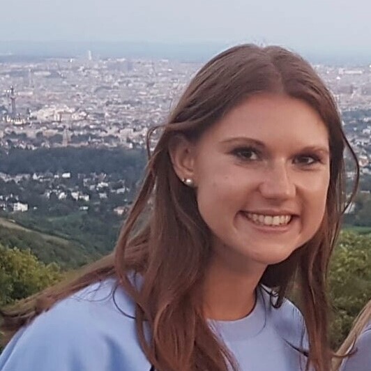 Victoria Meil trägt einen hellblauen Pullover und hat lange braune Haare. Sie steht auf einer Erhöhung, im Hintergrund sieht man das Panorama einer großen Stadt.