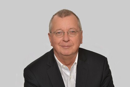 Portrait von Martin Weber. Er trägt ein weißes Hemd sowie einen schwarzen Blazer und sitzt lächelnd vor einer grauen Wand.