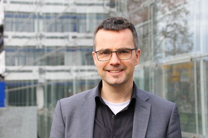 Portrait von Marc Debus. Er trägt ein schwarzes Hemd sowie einen grauen Blazer und steht vor dem A5-Gebäude.