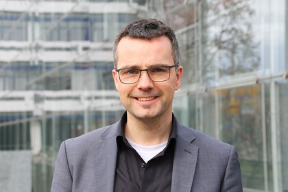 Portrait von Marc Debus. Er trägt ein schwarzes Hemd sowie einen grauen Blazer und steht vor dem A5-Gebäude.