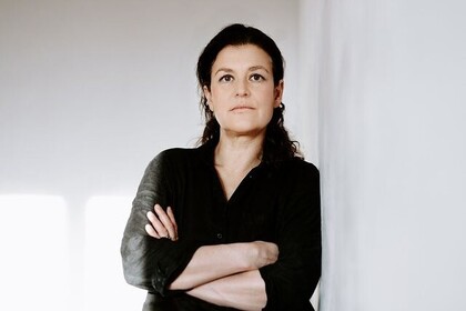 Porträt der Alumna Dr. Uta Meeder. Sie lehnt mit vor der Brust verschränkten Armen seitlich an einer hellen Wand. Sie blickt dabei mit ernstem Blick direkt in die Kamera. Sie trägt eine schwarze Bluse und das dunkle lange Haar zu einem Teil nach hinten gebunden. Link: Instagram Post Dr. Uta Meeder