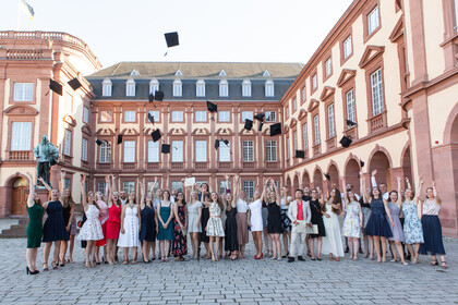 Mehrere Studierende stehen vor dem Mannheimer Schloss und werfen Absolventenhüte in die Luft. 