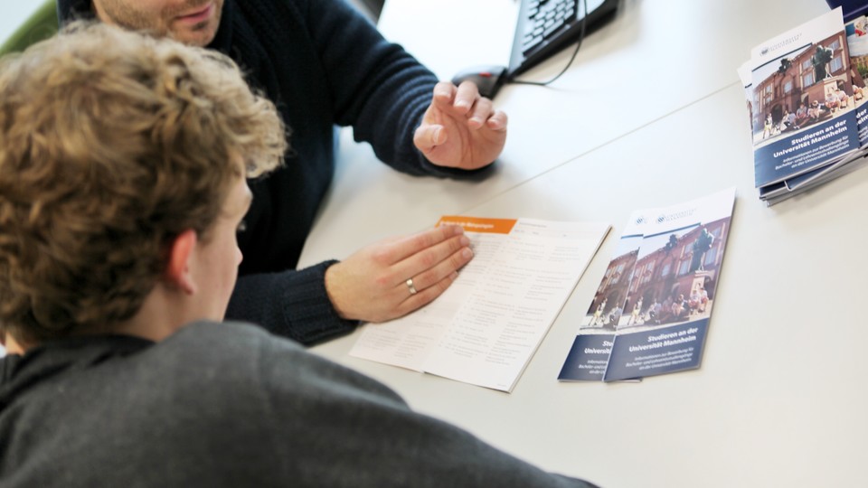 Ein Mitarbeiter berät einen Studenten mit einem Flyer zum Thema "Studieren an der Universität Mannheim".