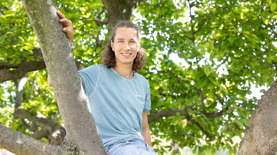 Luca Knörzer. Er sitzt lächelnd auf einem Ast. Er trägt ein hellblaues T-Shirt und seine Haare zur Hälfte hochgesteckt.