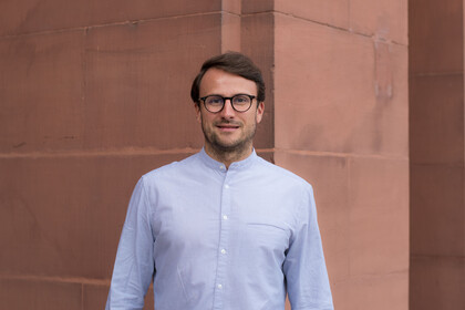 Portrait of Dr. Maximilian Beichert. He is wearing a white shirt and standing at an entrance to Mannheim Palace.