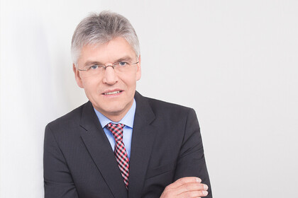 Portrait von Christoph Spengel. Er trägt ein schwarzes Jackett und lehnt an eine weiße Wand.