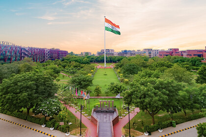 Im Hintergrund sieht man eine Indien-Flagge. Sie steht in einem Park, der von Gebäuden umgeben ist. Im Vordergrund steht silbern "JGU"