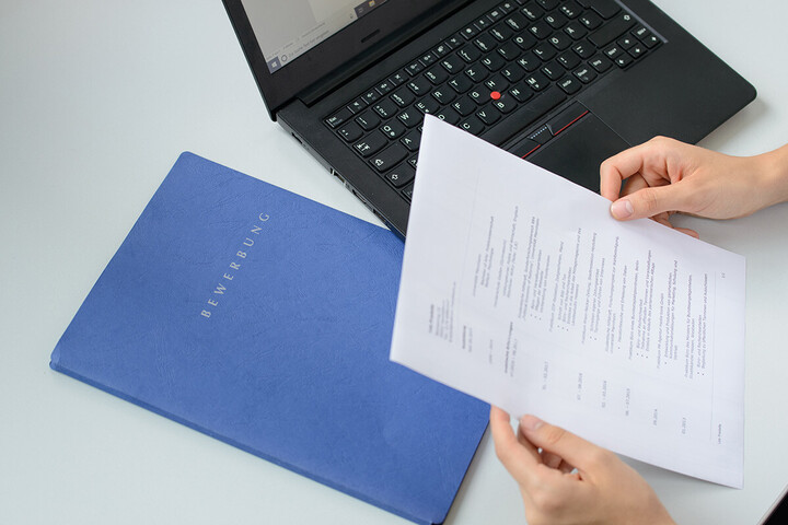 Eine blaue Mappe liegt auf dem Tisch, auf der Bewerbung geschrieben steht. Zu sehen sind die Hände zweier Personen. Eine hält eine Broschüre in den Händen, die andere einen Kugelschreiber und einen Schreibblock.