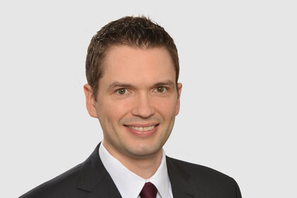 Portrait von Florian Kraus. Er trägt ein weißes Hemd, eine rote Krawatte sowie einen schwarzen Blazer und steht lächelnd vor einer weißen Wand.