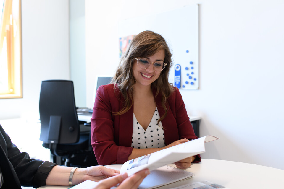 Eine Person sitzt gemeinsam mit einer anderen Person, die Dokumente in der Hand hält, an einem Tisch. Sie blickt lächelnd auf die Dokumente.