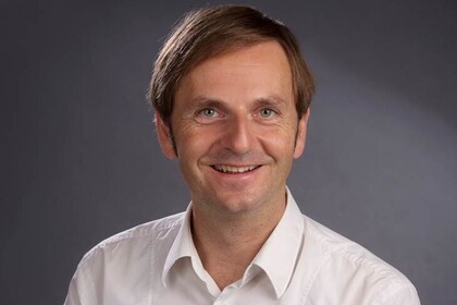Portrait von Oliver Dickhäuser. Er trägt ein weißes Hemd und steht vor einem grauen Hintergrund. 