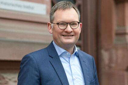 Portrait von Philipp Gassert. Er trägt ein hellblaues Hemd sowie einen dunkelnblauen Blazer und steht lächelnd vor einem Gebäude.