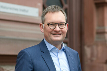Portrait von Philipp Gassert. Er trägt ein hellblaues Hemd sowie einen dunkelnblauen Blazer und steht lächelnd vor einem Gebäude.