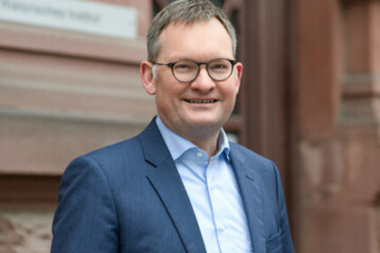 Portrait von Philipp Gassert. Er trägt ein hellblaues Hemd sowie einen dunkelnblauen Blazer und steht lächelnd vor einem Gebäude.