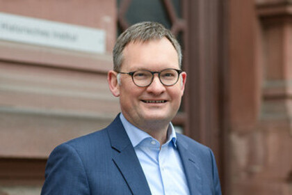 Portrait von Philipp Gassert. Er trägt ein hellblaues Hemd sowie einen dunkelnblauen Blazer und steht lächelnd vor einem Gebäude.
