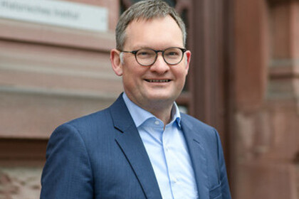 Portrait von Philipp Gassert. Er trägt ein hellblaues Hemd sowie einen dunkelnblauen Blazer und steht lächelnd vor einem Gebäude.