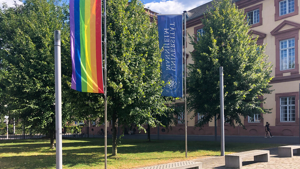 Regenbogenflagge und Flagge der Universität Mannheim wehen vor dem Ostflügel des Mannheimer Schlosses, Bäumen im Hintergrund.