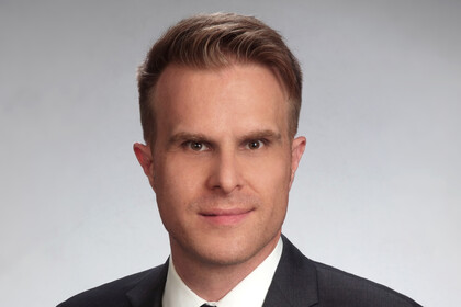 Portraitfoto von Prof. Dr. Florian Stahl vor einem grauen Hintergrund. Er trägt ein weißes Hemd, eine schwarze Krawatte und ein schwarzes Sakko.