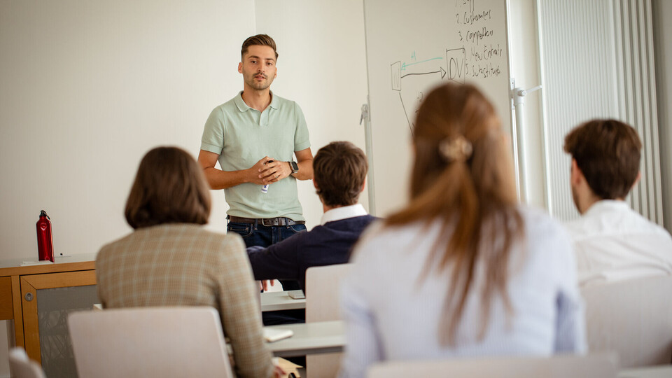 Junger Mann steht vor einer Klasse von Doktorand*innen