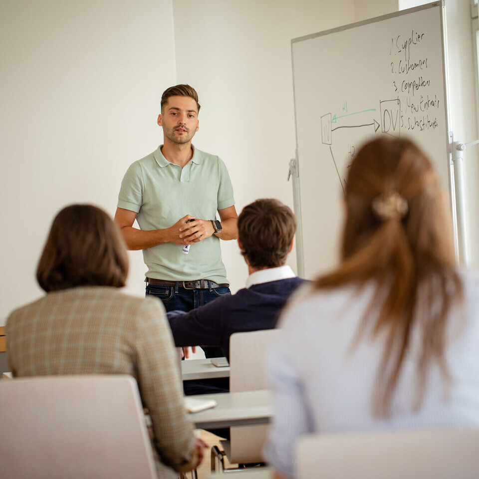 Junger Mann steht vor einer Klasse von Doktorand*innen