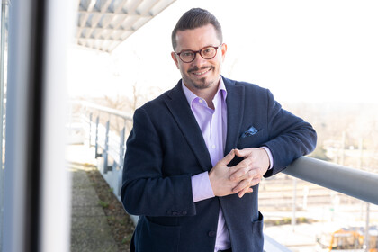 Eine lächelnde Person trägt ein lilafarbenes Hemd sowie einen dunkelblauen Blazer und lehnt an ein Geländer. Die Person heißt Nicolas Ziebarth.