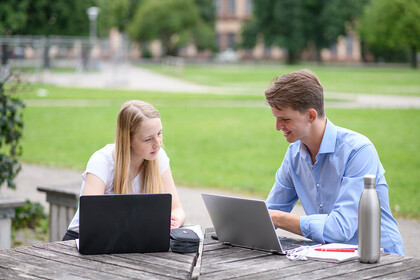 Zwei Studierende sitzen an einem Tisch im Freien auf dem Gelände der Universität Mannheim. Sie sitzen vor ihren Laptops und unterhalten sich.