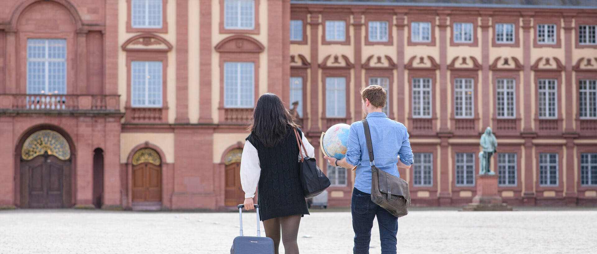 Zwei Studierende laufen auf das Schloss zu. Eine Person zieht einen kleinen Rollkoffer nach sich, die andere hält einen Globus in der Hand.