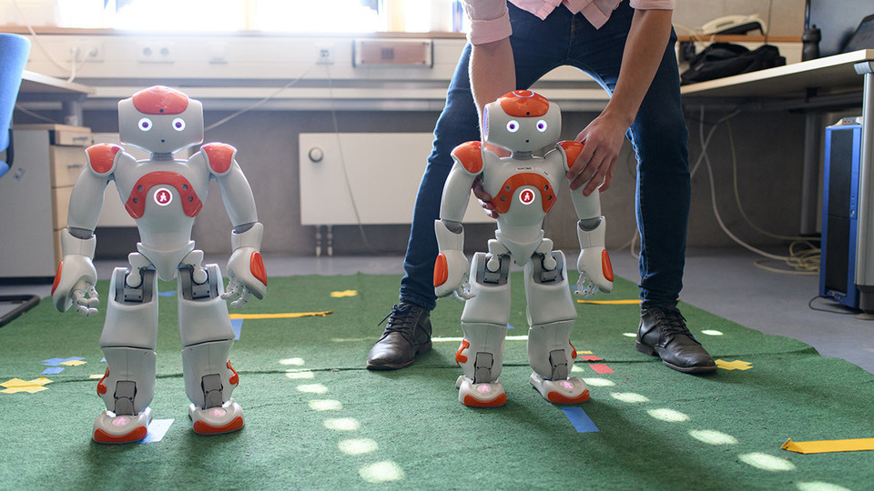 zwei kleine Roboter in einem Raum, der rechte wird von einem Mann geführt