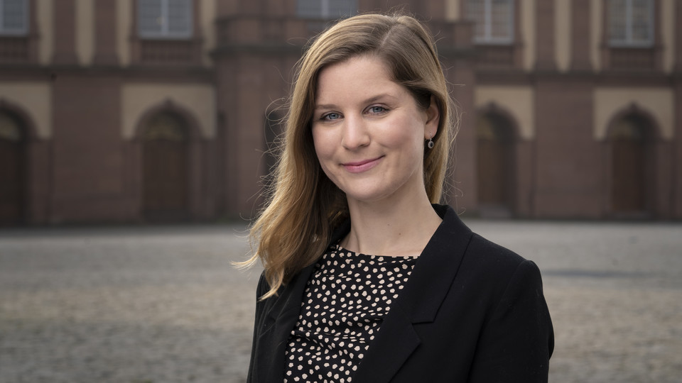 Lea Oberländer hat blonde lange Haare und trägt ein gemustertes Oberteil und einen dunklen Blazer. Im Hintergrund ist das Schloss zu sehen.