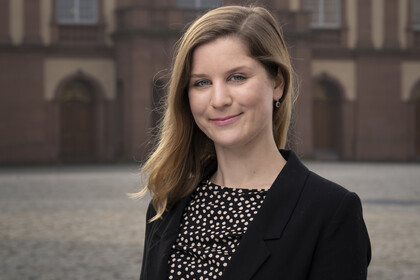 Lea Oberländer hat blonde lange Haare und trägt ein gemustertes Oberteil und einen dunklen Blazer. Im Hintergrund ist das Schloss zu sehen.