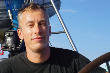 Stefan Kluge im Sonnenschein auf einem Segelboot. Er trägt ein schwarzes T-Shirt und eine Sonnenrille auf dem Kopf.