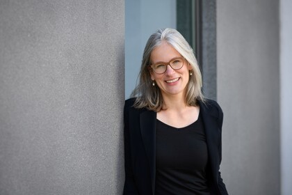Gesundheitspsychologin Prof. Dr. Jutta Mata lehnt an eine Betonwand und lächelt. Sie trägt einen schwarzen Blazer und ein schwarzes T-Shirt.