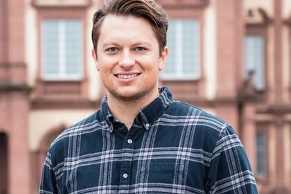 Gordon Weuste hat kurze braune Haare. Er trägt ein dunkelblau-kariertes Hemd und lächelt in die Kamera