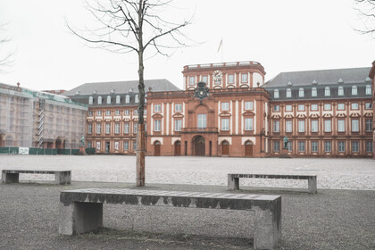 Blick auf den Mittelbau der Universität Mannheim. Der Himmel ist grau und im Vordergrund sind Sitzbänke zu sehen