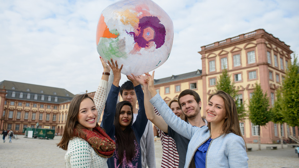 Eine Gruppe Studierender hält eine Weltkugel in die Luft. Sie stehen auf dem Gelände der Unversität Mannheim und lächeln in die Kamera.