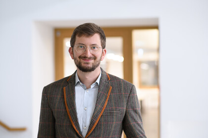 Philipp Müller trägt ein weißes Hemd sowie einen braun, orange karrierten Blazer. Er steht vor einer Glastüre.