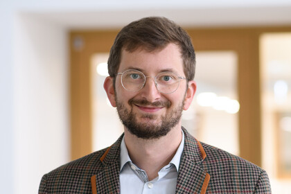 Portrait von Philipp Müller. Er trägt ein weißes Hemd sowie einen braun orange karrierten Blazer und steht in einem gebäude vor einer Glastüre.