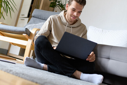 Studierender mit kurzen braunen Haaren und einem beigen Kapuzenpullover sitzt auf dem Boden mit einem Laptop auf dem Schoß.