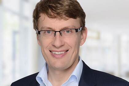 Portrait von Klaus Adam. Er trägt ein weißes Hemd sowie einen dunkelblauen Blazer und steht lächelnd vor einer Fensterfront.