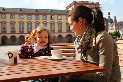 Frau mit kleinem Kind sitzt im Ehrenhof der Universität, das Kind isst eine Bretzel, die Frau trinkt Kaffee, sonniger Tag.