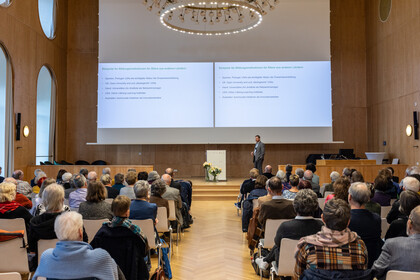Foto von der Jubiläumsveranstaltung in der Aula. Das Publikum hört dem Redner auf dem Podium zu. Hinter ihm ist eine Präsentation an die Wand projiziert.