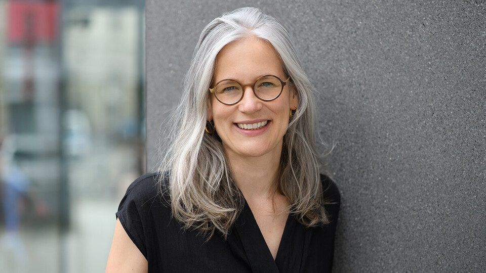 Porträt von Jutta Mata. Eine Frau mit langen grau-weißen Haaren, einer Brille und einer blauen Bluse.