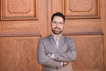 Portrait von Studierende*m Talha Güney (dunkle Haare, Hemd, grauer Anzug) vor einer Holztür.
