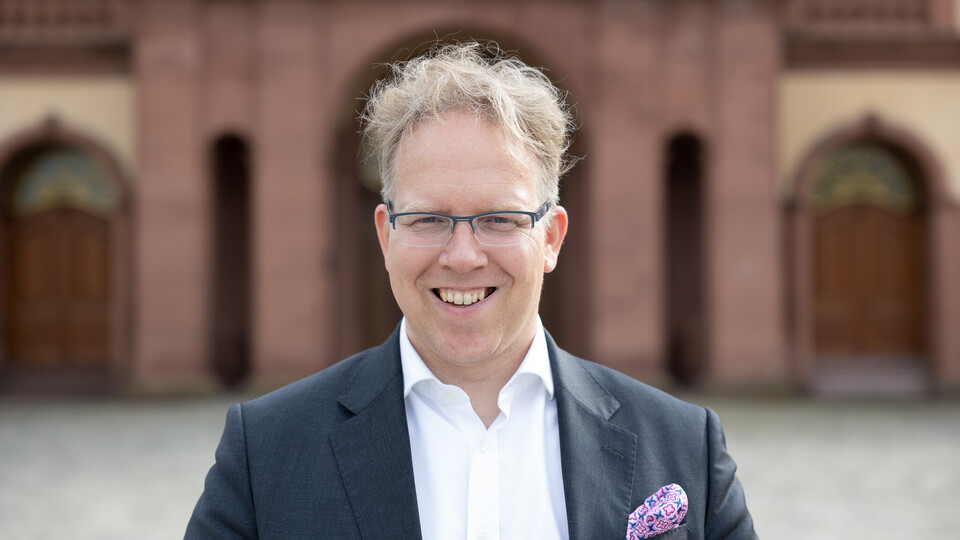 Portrait von Oliver Brand. Er trägt ein weißes Hemd sowie einen grauen Blazer und steht lächelnd auf dem Ehrenhof.