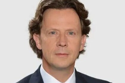 Michael Woywode hat leicht gewellte, etwas längere dunkle Haare. Er trägt ein helles Hemd, eine hellblaue Krawatte und ein blaues Jacket.
