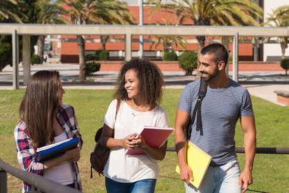 Hintergrund: grüne Wiese und ein Gebädue, im Vordergrund: drei Studenten (zwei Frauen, ein Mann), die mit Lernunterlagen lächelnd laufen