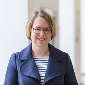 Ein Bild von Prof. Dr. Karina Karst in einem langen Flur der Universität. Sie hat blonde kürzere Haare, trägt eine Brille und lächelt. Sie trägt ein gestreiftes Oberteil mit einem dunkelblauen Blazer darüber.