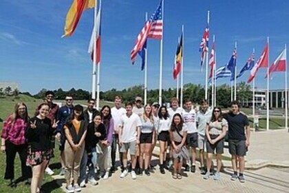 Gruppe junger Menschen posiert vor internationalen Flaggen im Freien bei sonnigem Wetter.