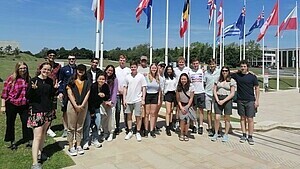 Gruppe junger Menschen posiert vor internationalen Flaggen im Freien bei sonnigem Wetter.