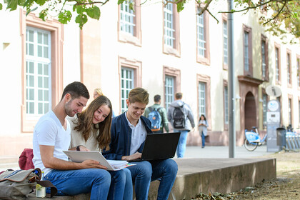 Drei Personen sitzen nebeinander auf einer Steinmauer. Zwei Personen schauen in einen Ordner, während die andere Person auf ihren Laptop blickt.